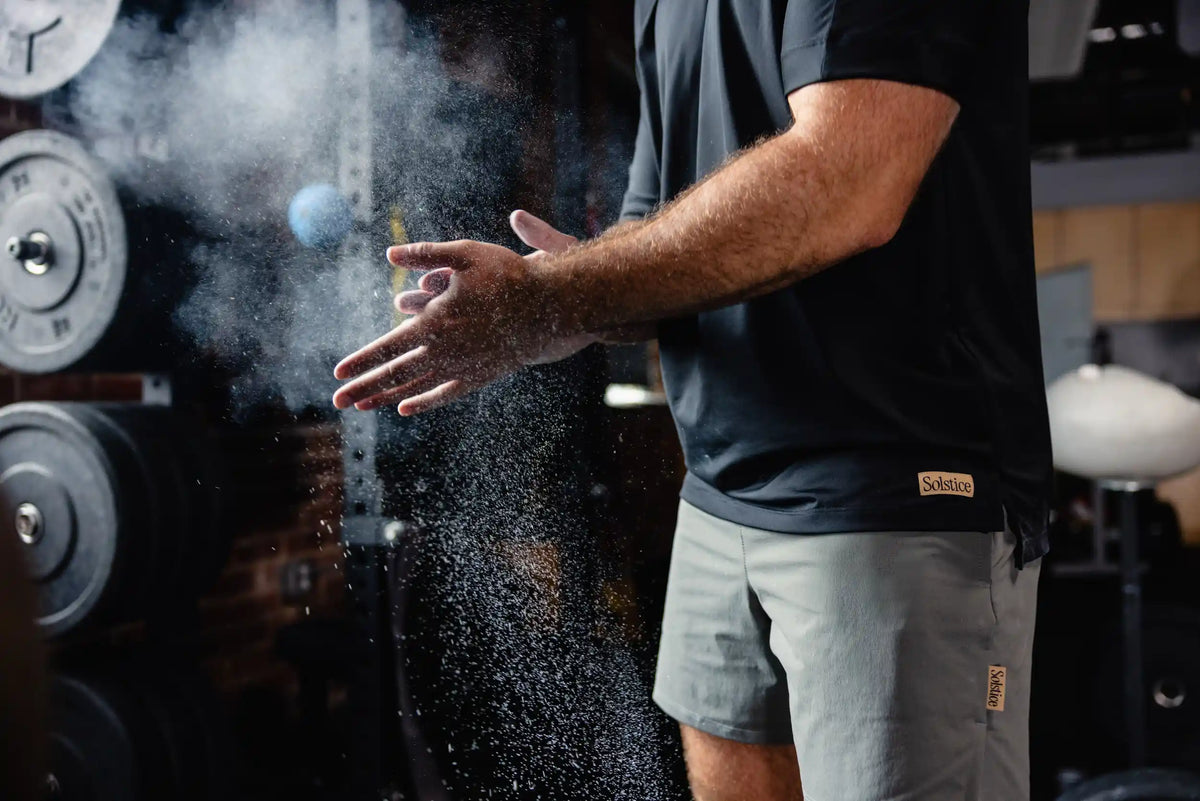 Athlete wearing a dark Solstice tee and grey shorts, chalking their hands in a gym.
